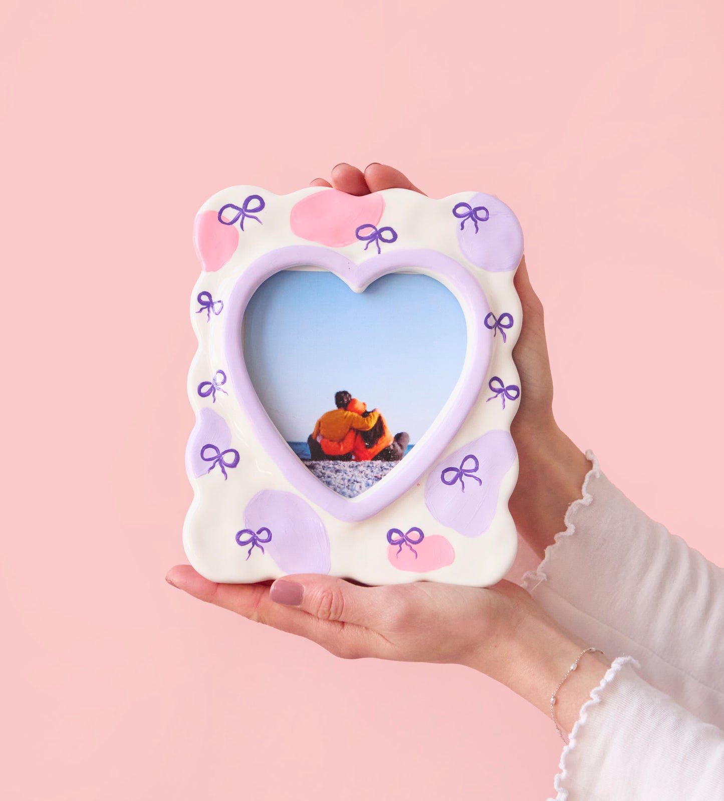 Heart-shaped photo frame with pink and purple decorative bows held in hands on a pastel pink background.