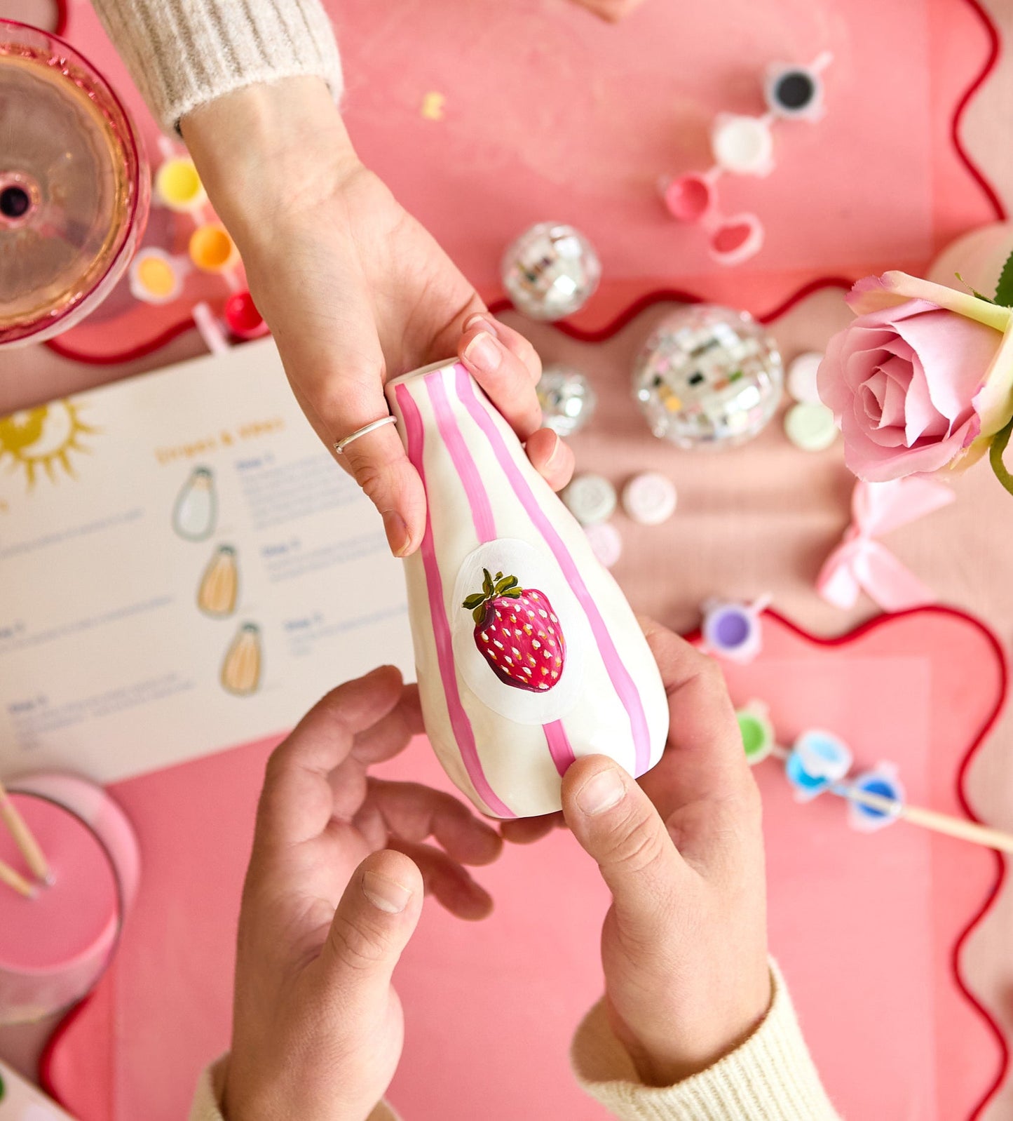 Person painting a white ceramic mug with colorful heart designs using a fine brush on a pink tabletop setting.
