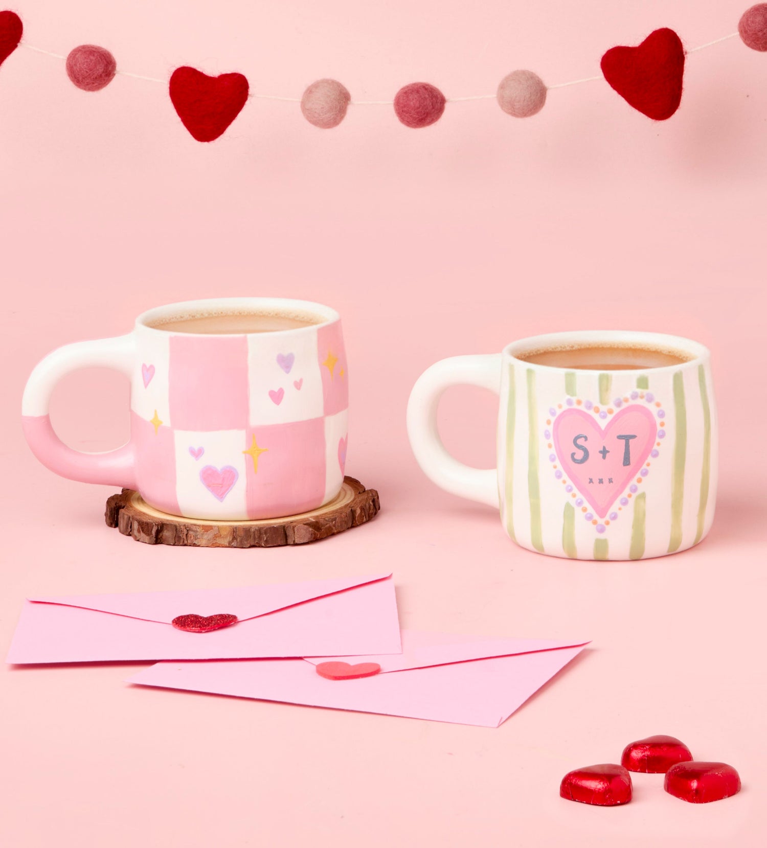 Two ceramic mugs with heart designs and handles on a pink table, alongside pink envelopes and red heart decorations.
