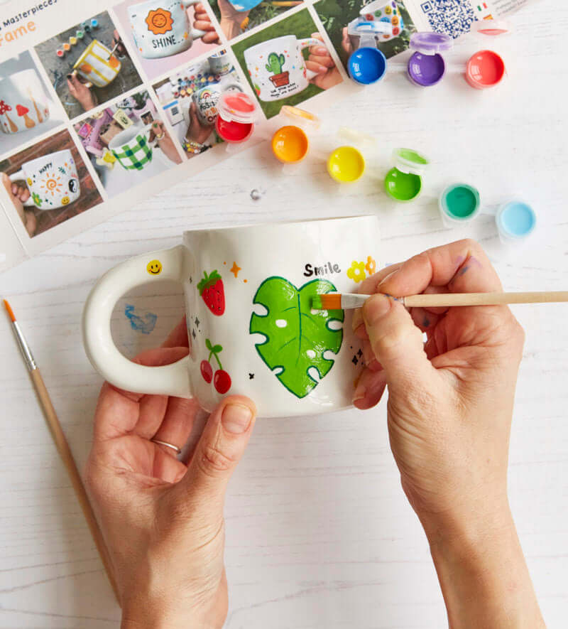Paint-a-mug kit with two custom mugs, vibrant ceramic paints, various paintbrushes, and step-by-step design inspiration guide.