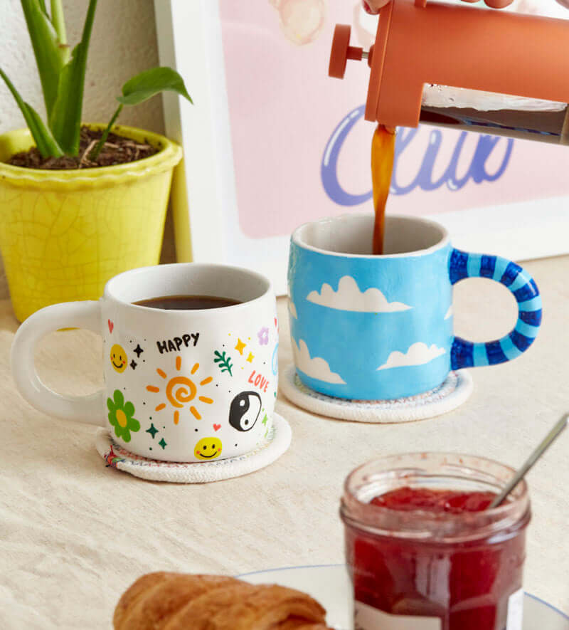 Hand painting a white ceramic mug with green checkered pattern using a thin brush for a creative DIY project.