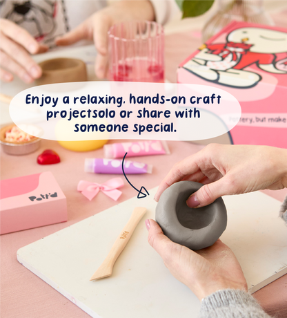 Hands shaping gray clay on a pink table with Pott'd crafting tools, wooden knife, and colorful packaging in the background.