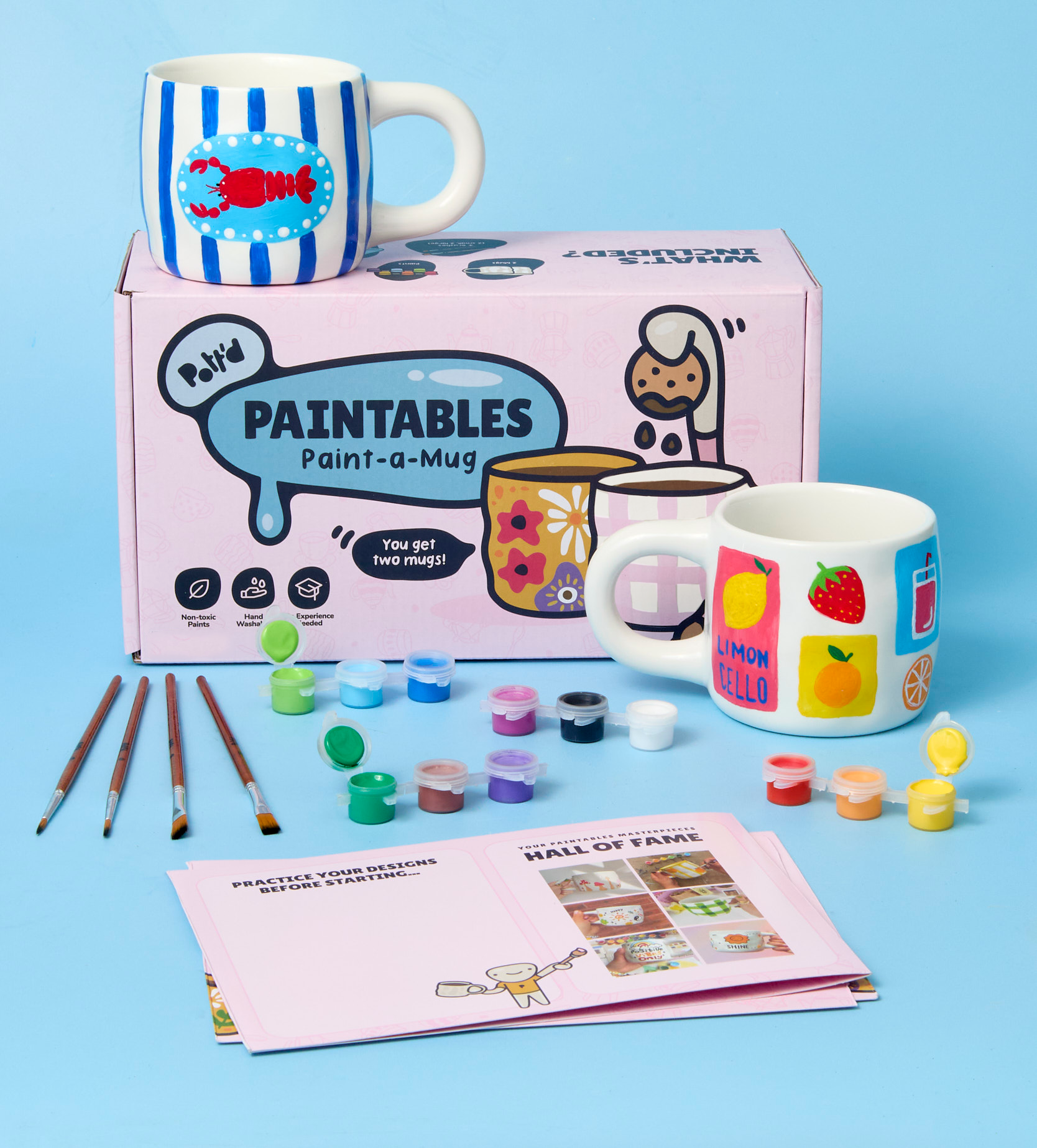 Ceramic mug painting kit with brushes, paints, and two customizable mugs with colorful designs, placed on a tabletop.
